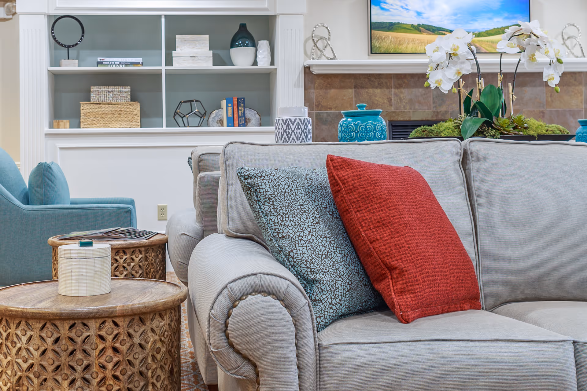 A cozy living room area featuring a light gray sofa with a red and a patterned blue cushion, a blue armchair, two wooden side tables with decorative items, built-in white shelves with books and decor, and a fireplace with a landscape painting above it and white orchids on the mantel.