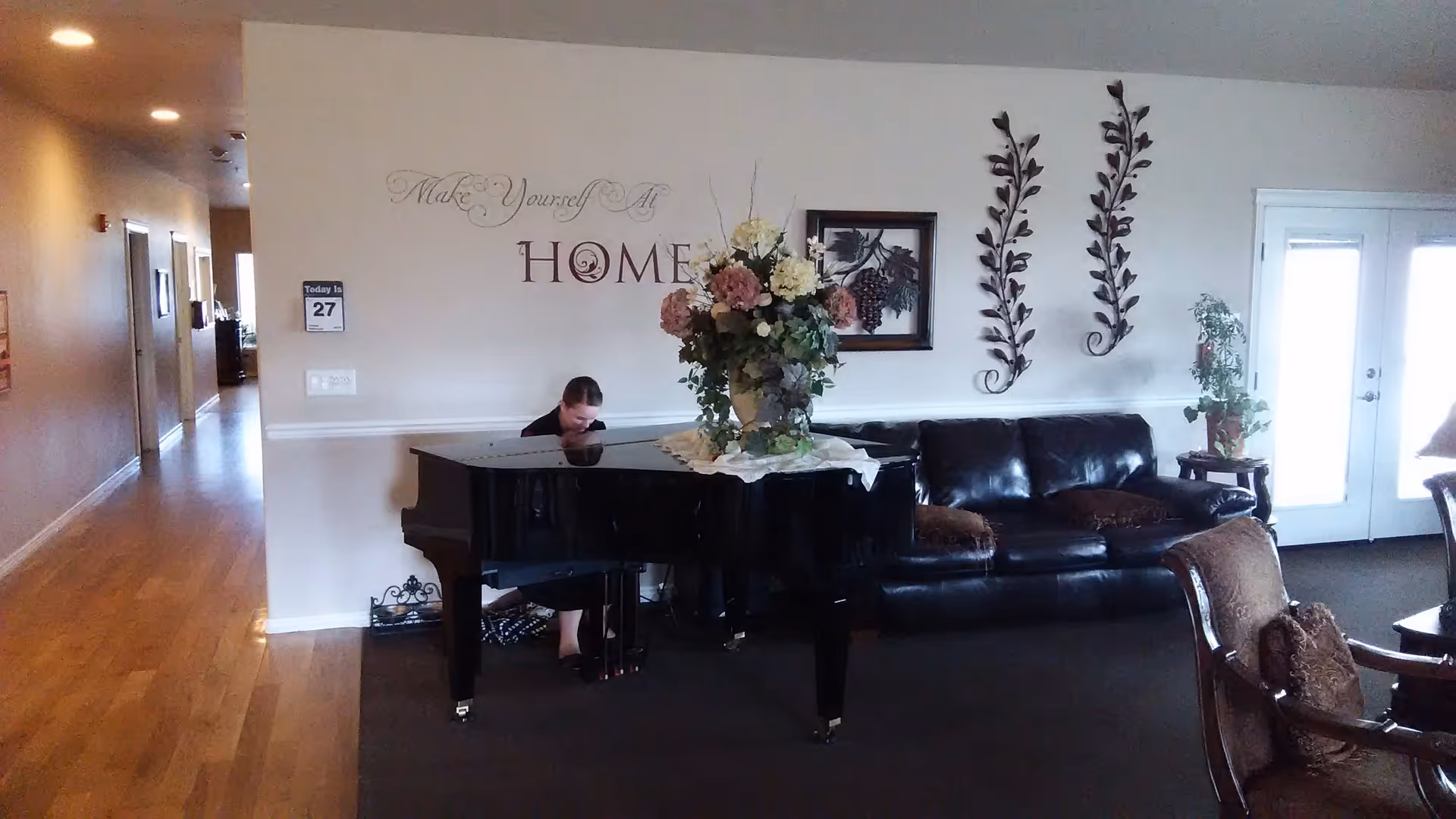 A cozy assisted living common area with a black grand piano adorned with a large floral arrangement. A person is seated at the piano playing. Behind the piano is a black leather couch with decorative pillows, wall art including a framed picture and metal leaf sculptures, and a wall decal that reads 'Make Yourself At HOME'. To the right, there are glass double doors letting in natural light. The floor is a combination of carpet and wood, with a hallway visible to the left.