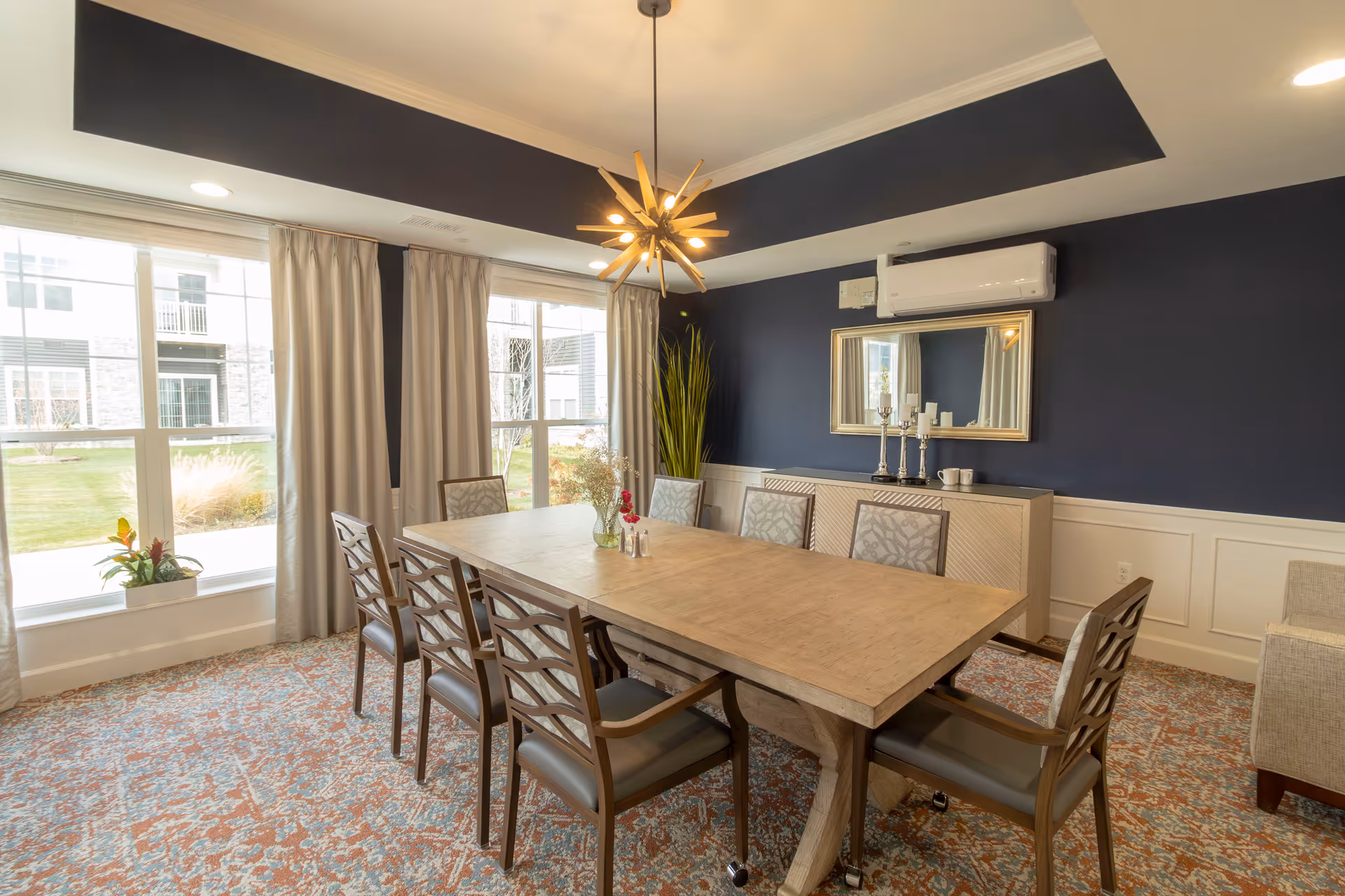 A well-lit dining room with a large wooden table surrounded by eight chairs. The room features a patterned carpet, dark blue walls with white wainscoting, a modern chandelier hanging from the ceiling, a large mirror on the wall, and a sideboard with decorative items. Large windows with beige curtains allow natural light to enter, showing a view of the outdoor greenery.