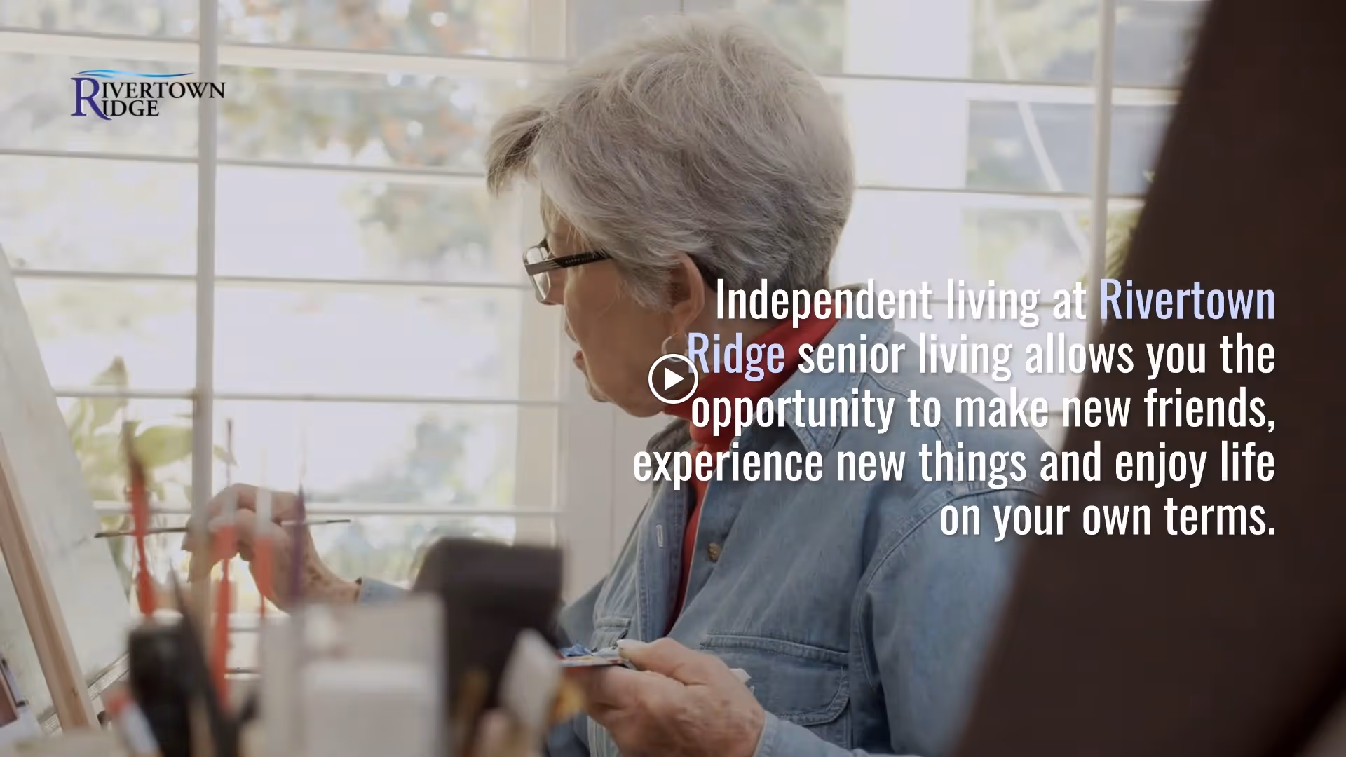 An elderly woman with gray hair and glasses is painting on a canvas near a window with blinds. She is wearing a denim jacket and appears focused on her artwork. The image includes text promoting independent living at Rivertown Ridge senior living.