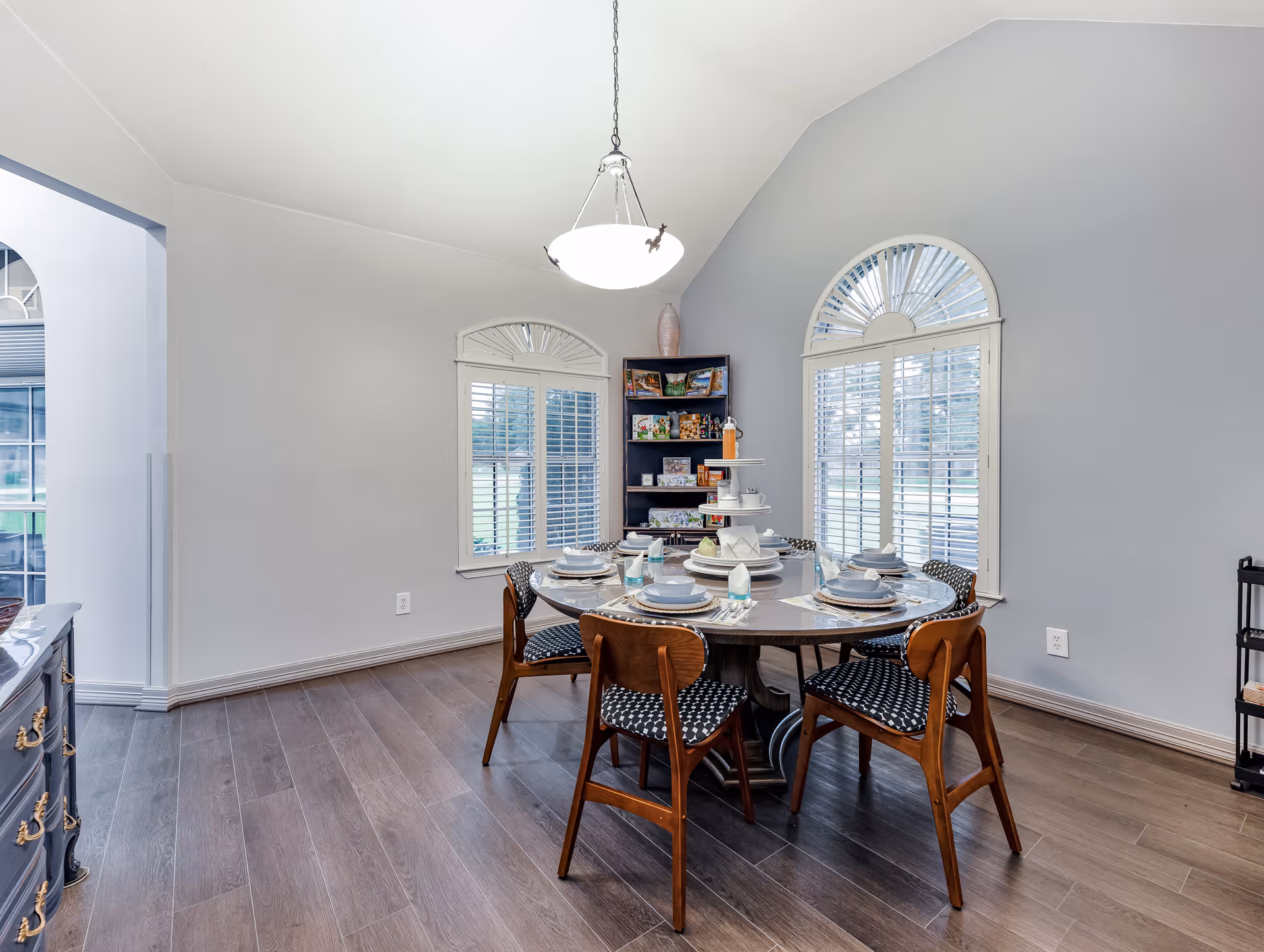 A dining room with a round table set for six people with plates, bowls, glasses, and cutlery. The room has two large arched windows with white shutters, light gray walls, and wood flooring. A hanging light fixture is above the table, and a corner shelf with various items is visible in the background.