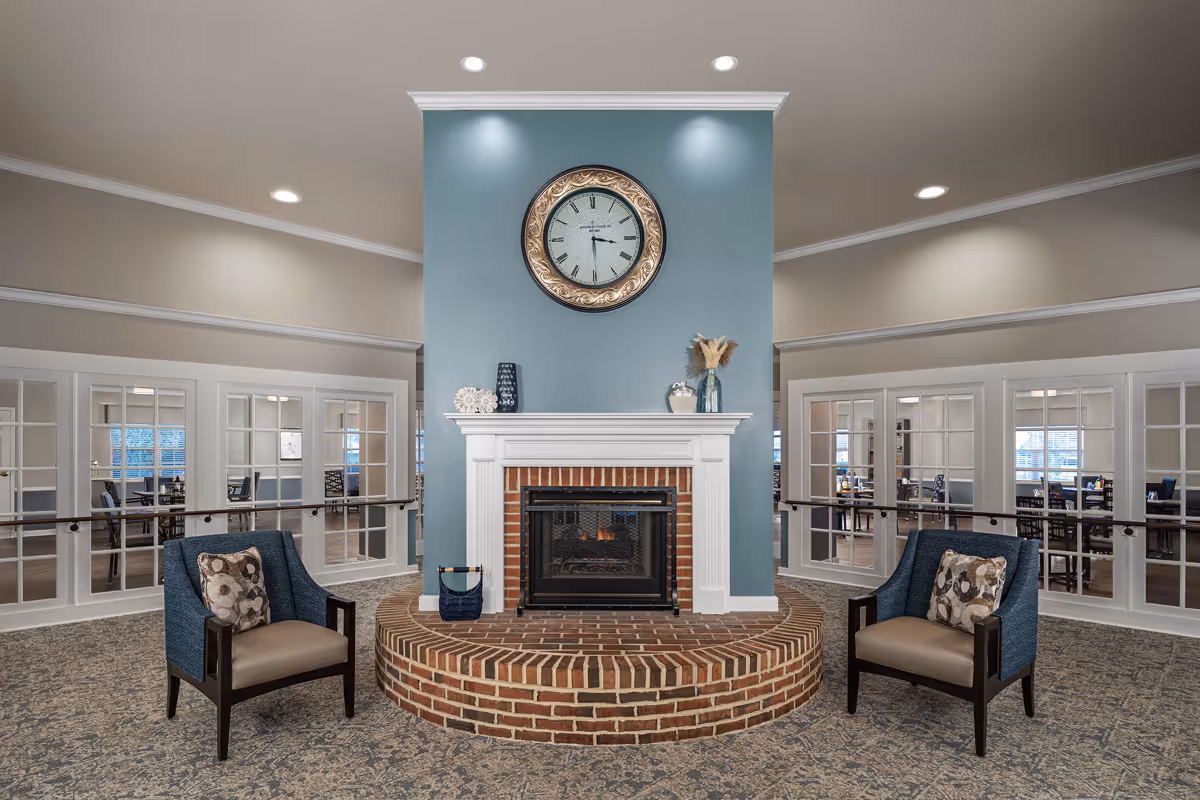 A cozy common area with a brick fireplace centered on a blue accent wall. Above the fireplace is a large round clock with a decorative frame. Two upholstered armchairs with patterned cushions are placed symmetrically on either side of the fireplace. Behind the fireplace, there are glass-paneled double doors leading to a dining area with tables and chairs. The room has recessed ceiling lights and a patterned carpet.