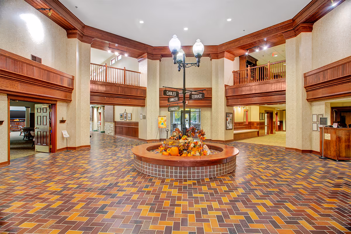 Spacious interior lobby area with a decorative round planter filled with autumn-themed decorations in the center. The floor is covered with multicolored brick tiles arranged in a herringbone pattern. The walls and ceiling feature wood paneling and trim, with a balcony railing visible on the upper level. A vintage-style street lamp post with directional signs is placed in the middle of the planter. Multiple doorways and corridors lead to other parts of the building.