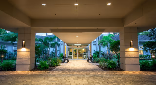 Covered porte-cochère leading to the glass entrance of a senior living building, flanked by landscaped beds and benches.