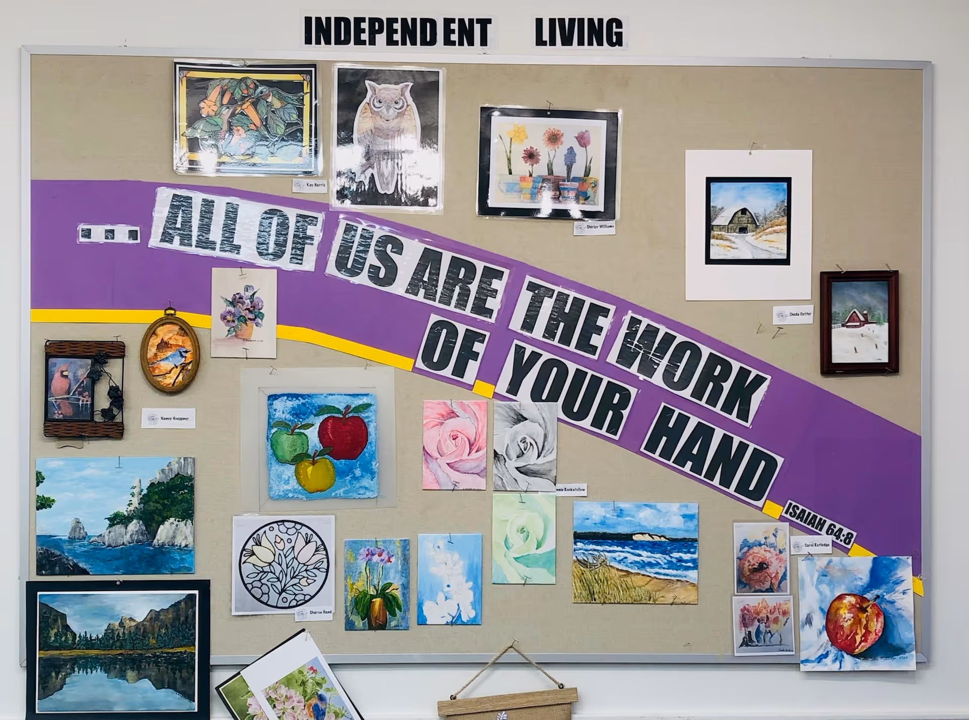 A bulletin board titled 'INDEPENDENT LIVING' displaying various colorful paintings and drawings including flowers, birds, apples, landscapes, and an owl. A purple and yellow banner across the board reads '...ALL OF US ARE THE WORK OF YOUR HAND ISAIAH 64:8'.