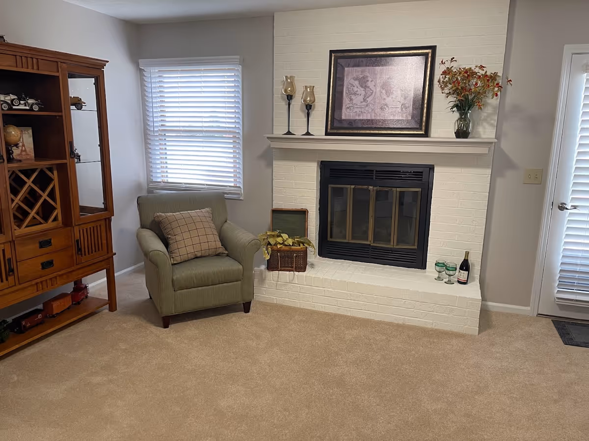 A cozy living room corner featuring a green armchair with a plaid cushion, a white brick fireplace with a black metal screen, and a wooden cabinet with shelves and drawers. On the fireplace mantel are two candle holders, a framed map, and a vase with flowers. There are also three green glasses and a bottle of wine on the hearth. A window with white blinds and a glass door with blinds are visible in the background.