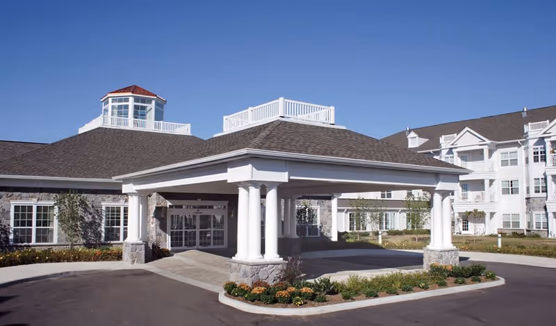Exterior view of a senior living facility building with a covered entrance supported by white columns, stone accents, and multiple windows under a clear blue sky.