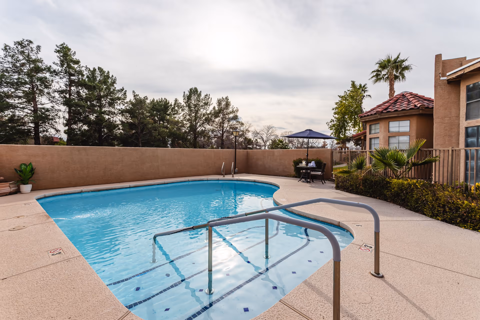 Outdoor swimming pool with clear blue water, metal handrails, and steps leading into the pool. Surrounding the pool is a beige textured deck with a small table and chairs under a blue umbrella near a tan stucco building with red tile roofing. Trees and shrubs are visible beyond a tan privacy wall under a cloudy sky.