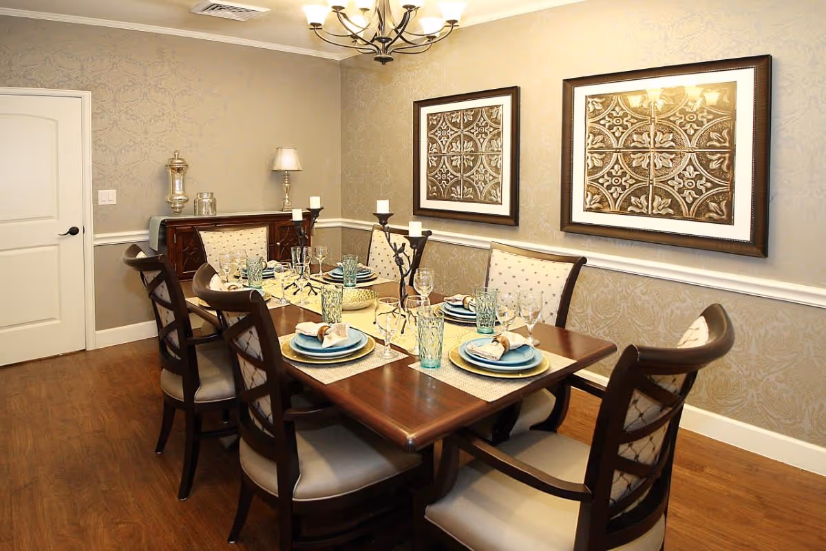 A formal dining room with a wooden dining table set for six people. The table is adorned with blue plates, beige napkins, clear drinking glasses, and decorative candle holders. The room features beige patterned wallpaper, two framed decorative artworks on the wall, a chandelier overhead, and a wooden sideboard against the back wall.