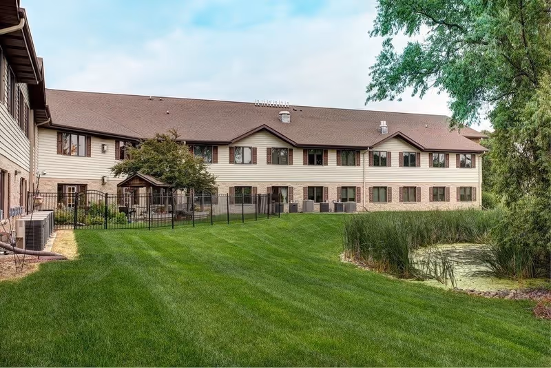 Exterior view of a two-story senior living facility building with beige siding and brown roof. The building is surrounded by a well-maintained green lawn, a small fenced garden area, and a pond with tall grass and trees nearby under a partly cloudy sky.