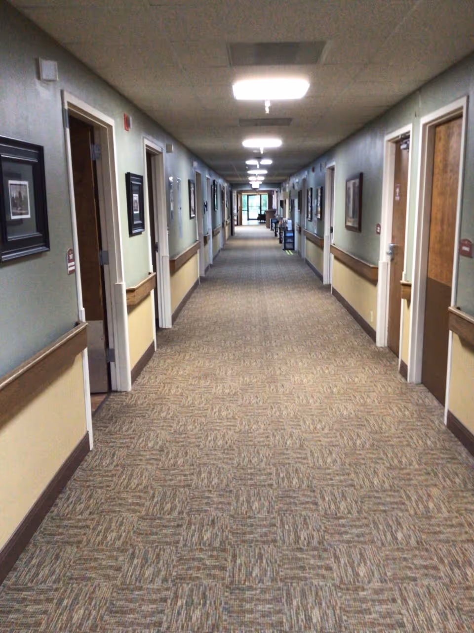 Long carpeted interior hallway with doors and framed pictures along both walls in a senior living facility.