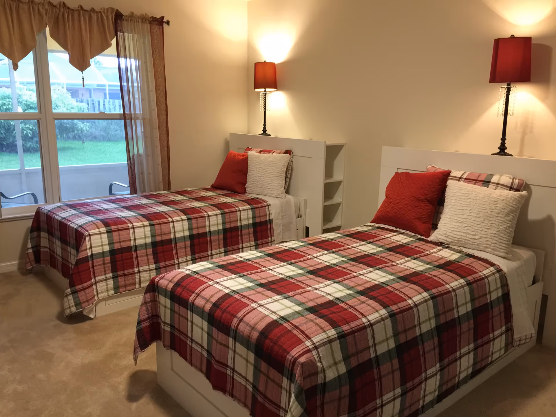 A bedroom with two single beds, each covered with a red, green, and white plaid bedspread. Each bed has two pillows, one red and one white. There are two matching lamps with red shades on the headboards, and a window with brown curtains showing a green lawn outside.