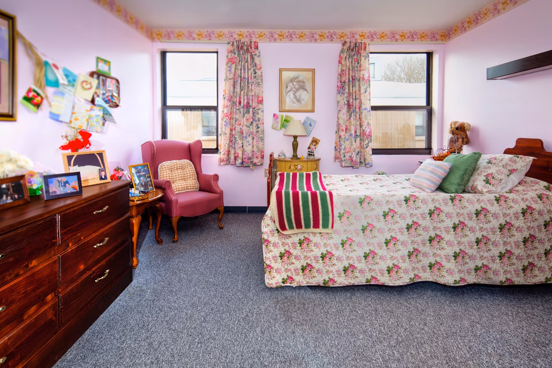 Sunlit bedroom with a floral bedspread, pink armchair, wooden dresser, and two windows.