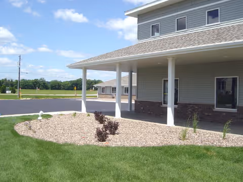 Covered entrance of a light-gray senior living building with columns, rock landscaping, and a grassy lawn under a blue sky.
