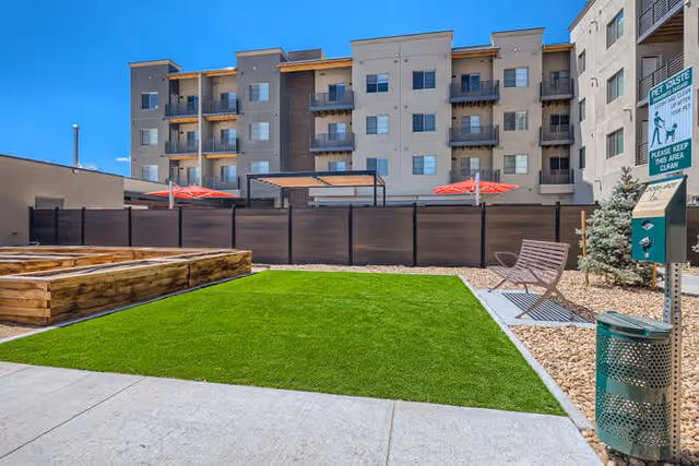 Outdoor area of a residential facility with a well-maintained green lawn, wooden raised garden beds, a bench, and a pet waste station. In the background, there is a multi-story apartment building with balconies and red umbrellas on the patio area.