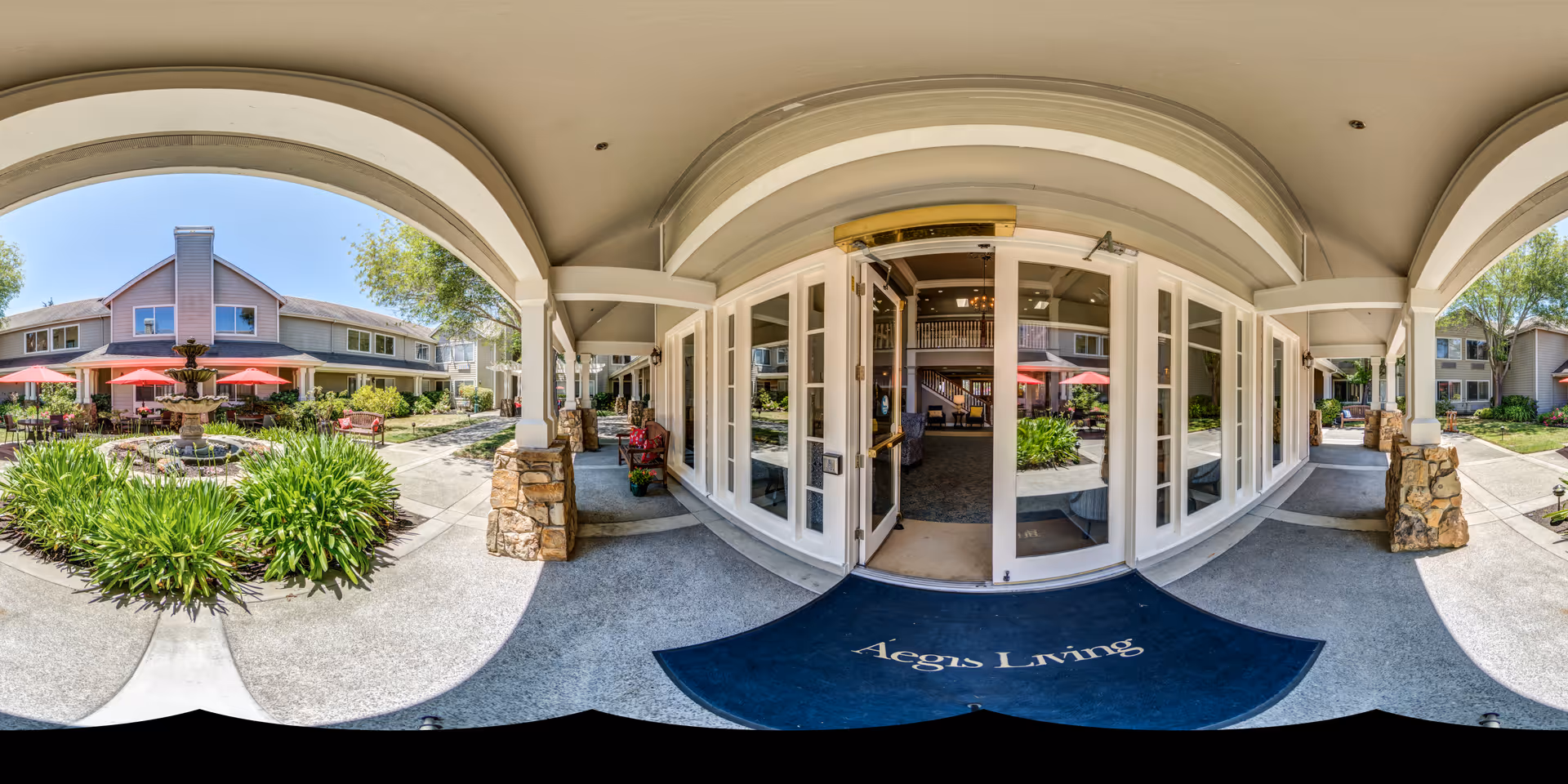 Outdoor courtyard area of Aegis Living Corte Madera featuring a covered walkway with stone pillars, glass doors leading inside, a fountain surrounded by greenery, and red umbrellas shading outdoor seating. The building has two stories with windows overlooking the courtyard.