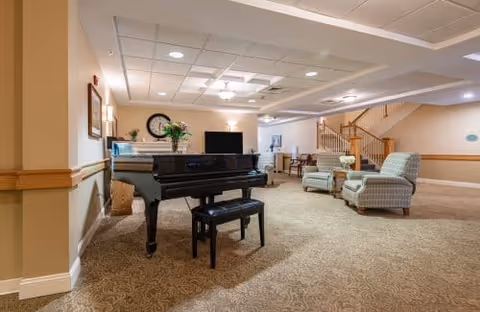 A comfortable senior living common room with a grand piano, upholstered armchairs, a TV, and a staircase in the background.