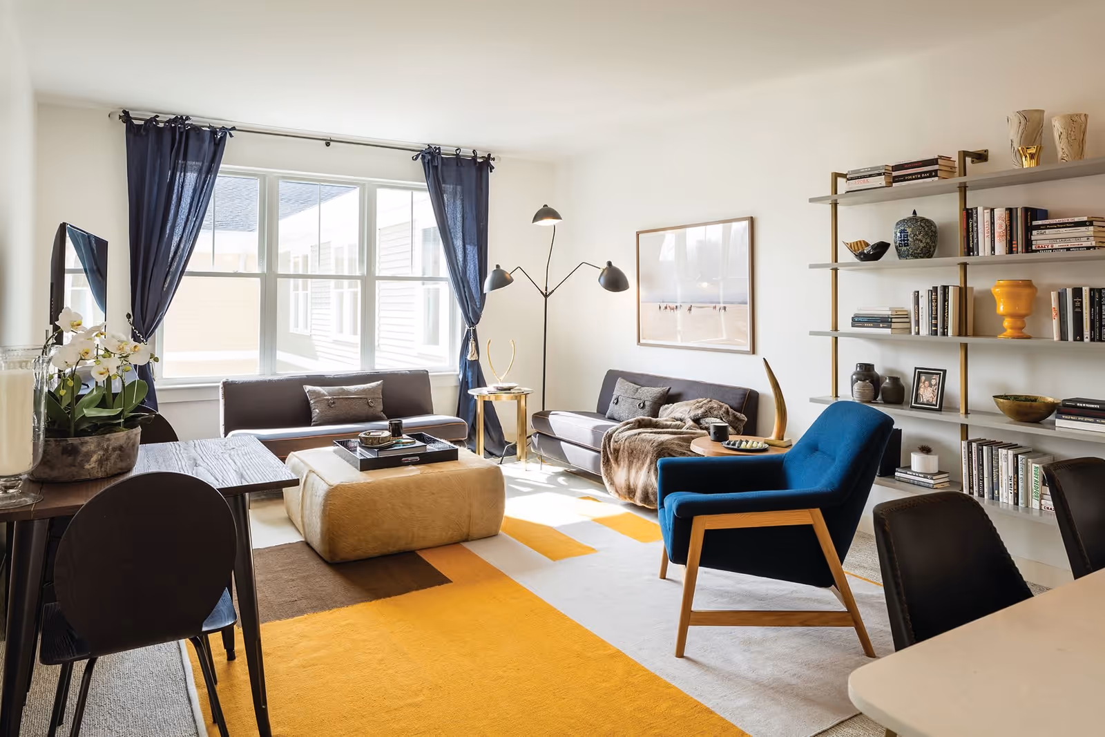 A bright and modern living room with large windows covered by dark blue curtains. The room features two gray sofas, a blue armchair, a beige ottoman, a wooden dining table with black chairs, a tall floor lamp, and a wall-mounted bookshelf filled with books and decorative items. The floor has a colorful yellow, brown, and white area rug.