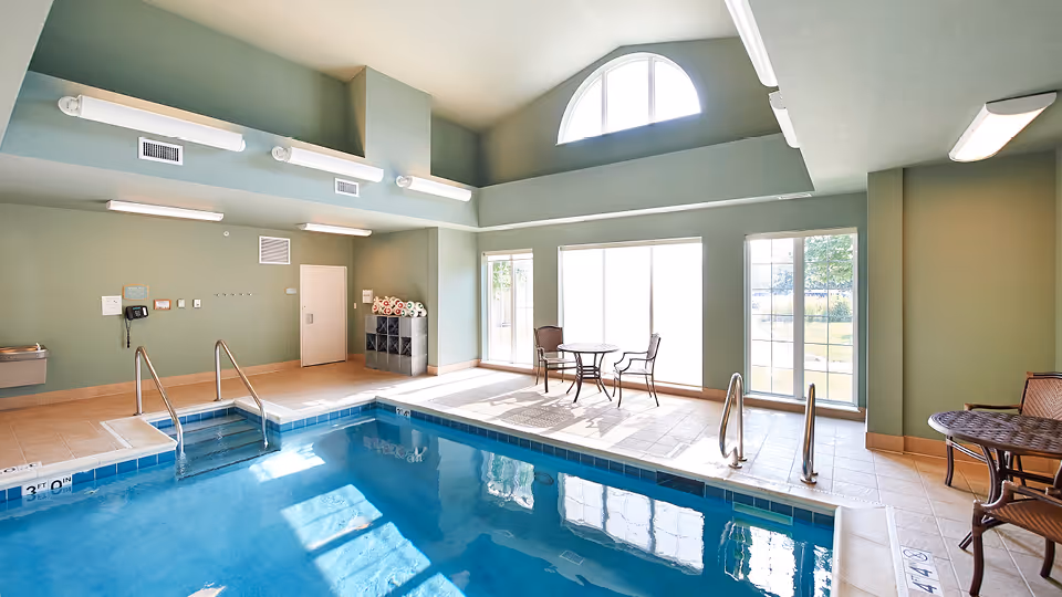 Indoor swimming pool area with clear blue water, tiled floor, metal handrails, and a few tables with chairs near large windows letting in natural light. The walls are painted light green and there is a high ceiling with fluorescent lights.