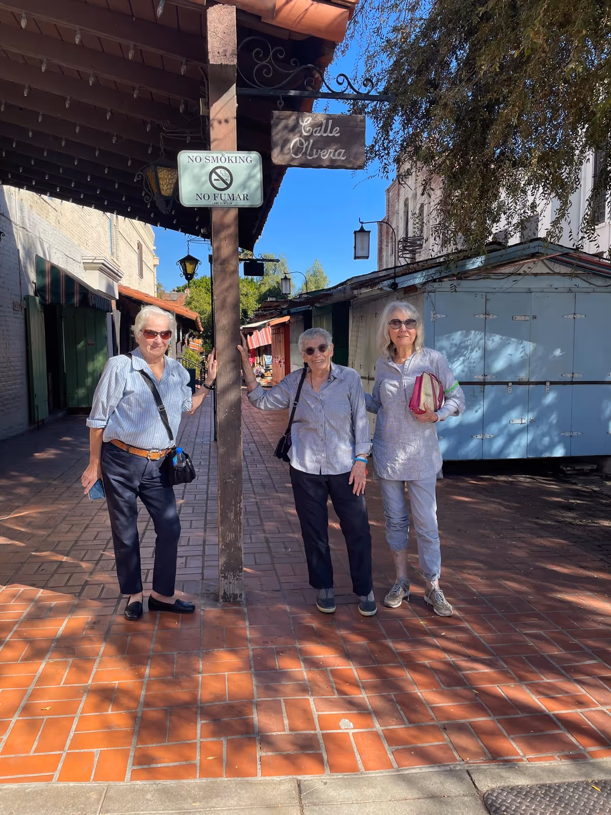 Three elderly women standing outdoors on a tiled walkway under a wooden awning. They are posing near a wooden post with a 'No Smoking / No Fumar' sign and a hanging sign that reads 'Calle Olvera'. The background shows closed market stalls and trees under a clear blue sky.