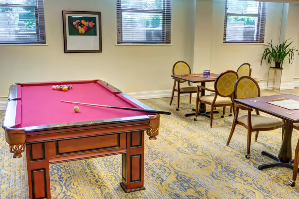 A recreation room with a red felt pool table set up for a game, two small tables with chairs around them, a framed picture of pool balls on the wall, three windows with blinds, and a potted plant on a stand in the corner.