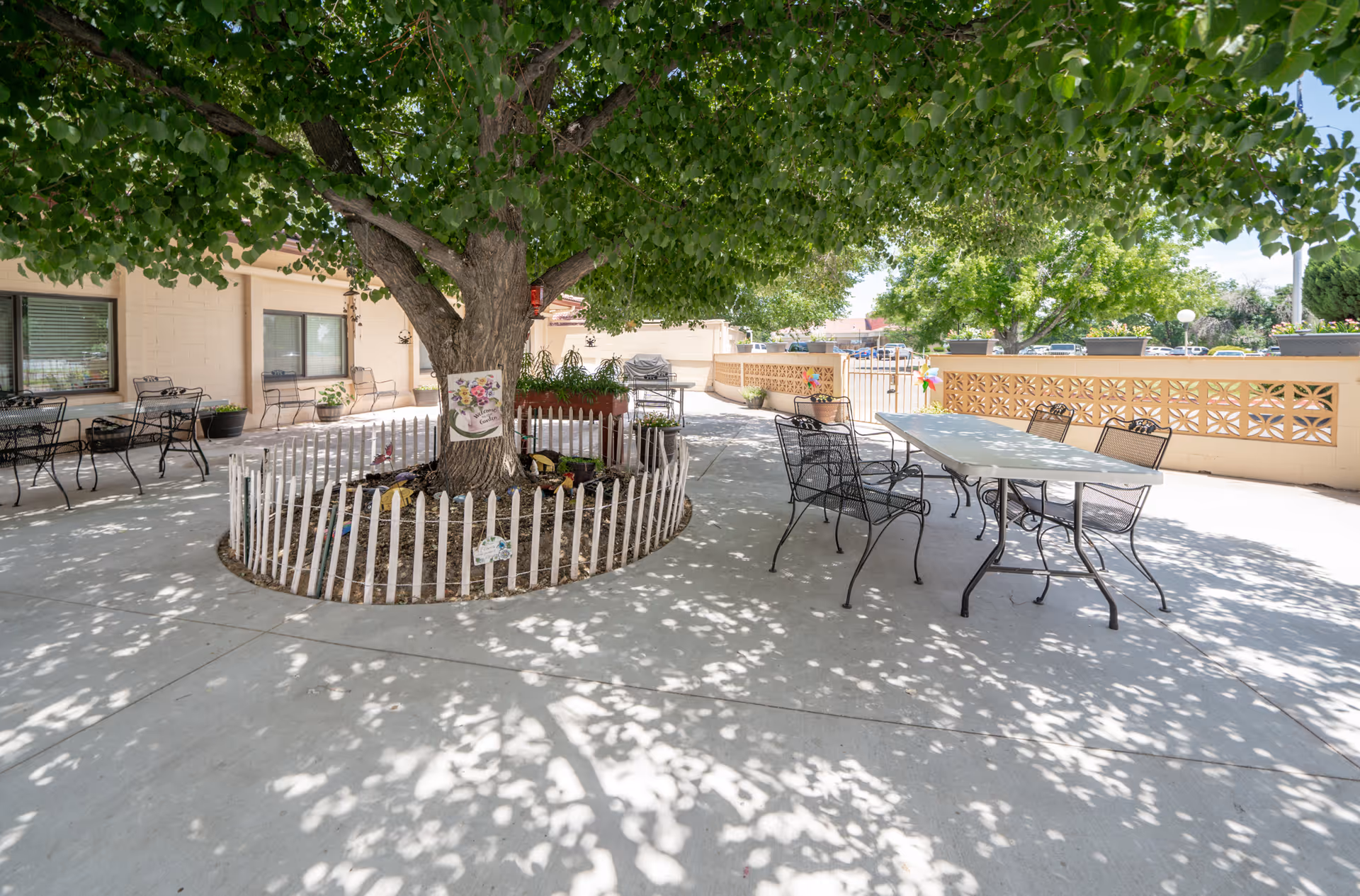 Outdoor patio area at La Villa Grande Care Center with a large tree surrounded by a white picket fence, several metal tables and chairs, and a beige building in the background. The area is shaded by the tree and there are potted plants and decorative items around.