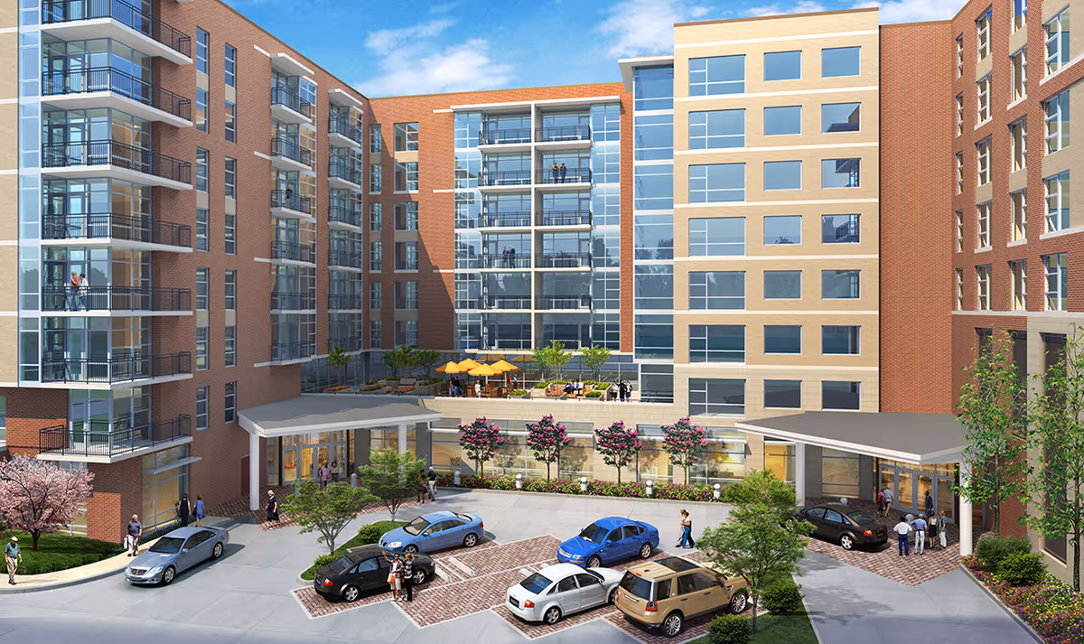 Exterior view of a multi-story senior living facility with large windows and balconies. There is a parking area with several cars and people walking near the entrance. The building is surrounded by landscaped greenery and trees, with a clear blue sky above.