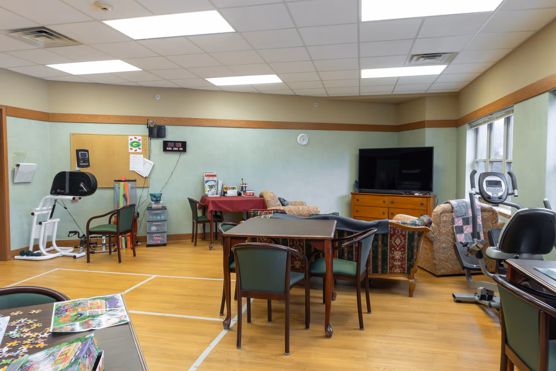 A senior living facility common room with exercise equipment, a television on a wooden dresser, several chairs and tables, a puzzle on one table, and a bulletin board on the wall. The room has wood flooring, light green walls with a wooden trim, and large windows letting in natural light.