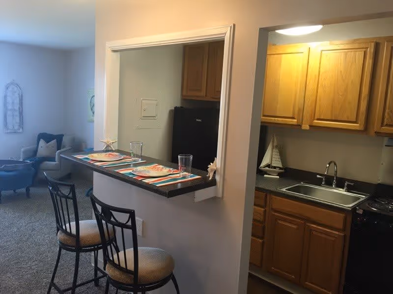 Interior view of a senior living facility showing a small kitchen area with wooden cabinets, a sink, and a stove. Adjacent to the kitchen is a breakfast bar with two chairs and place settings including plates, glasses, and placemats. In the background, there is a living room area with a blue armchair and a decorative wall piece.