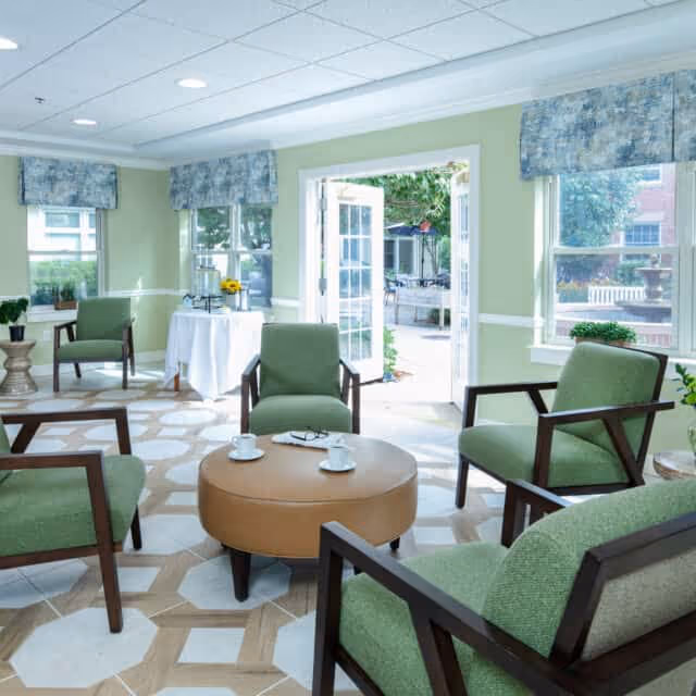 A bright and airy sitting area with four green upholstered armchairs arranged around a round tan ottoman with two white cups and saucers on it. The room has large windows with blue patterned valances and a door open to an outdoor patio area with tables and umbrellas. The floor has a geometric patterned tile design.