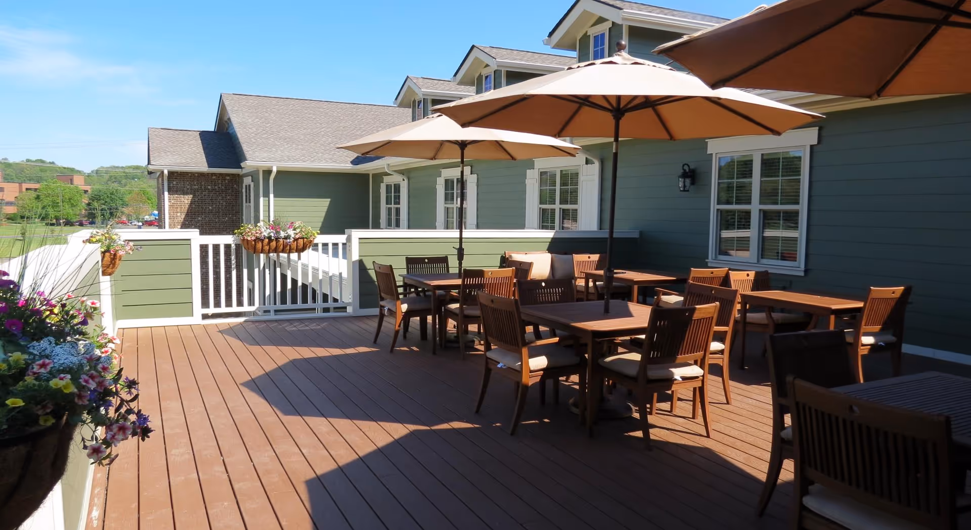 Outdoor patio area at Sycamore Springs Senior Living Community with wooden deck flooring, several wooden tables and chairs with cushions, and large beige umbrellas providing shade. The patio is adjacent to a green building with white-trimmed windows and flower pots hanging on the railing.