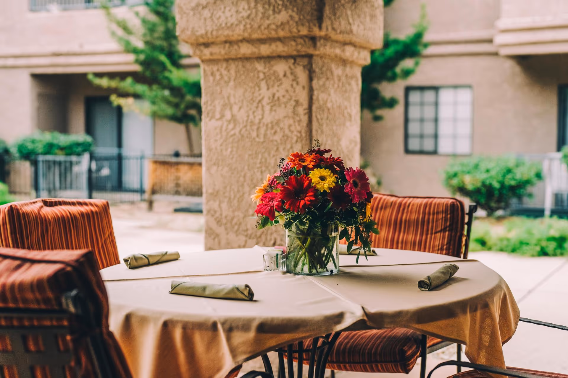 Round outdoor table on a covered patio set with a vase of colorful flowers and striped chairs in front of an apartment building.