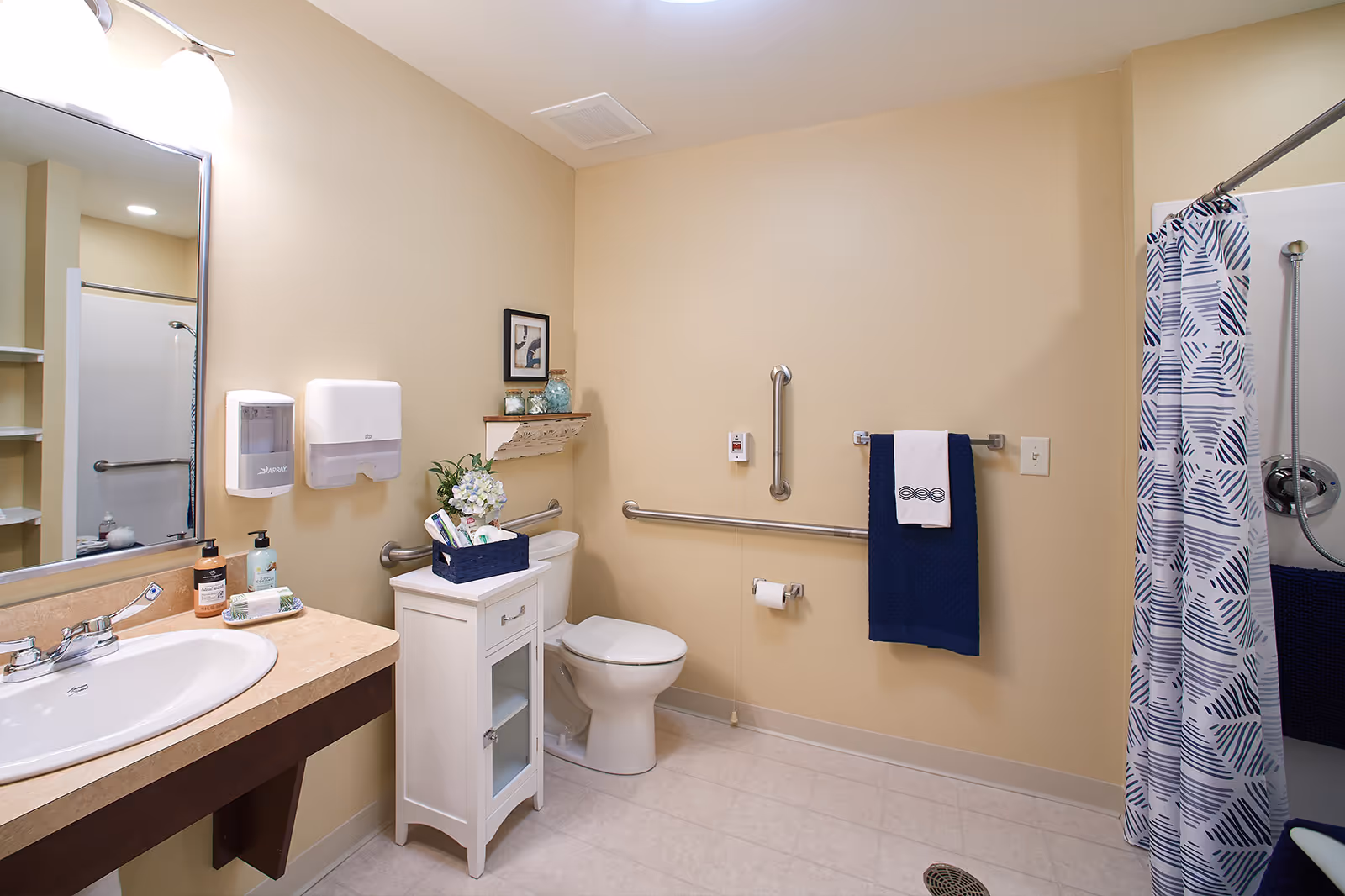 A clean and accessible bathroom with beige walls and tiled floor. It features a sink with a countertop and mirror, a white cabinet with decorative items and toiletries, a toilet with grab bars on the walls for support, a towel rack with a blue towel and a white hand towel, and a shower area with a patterned blue and white shower curtain and a handheld showerhead.