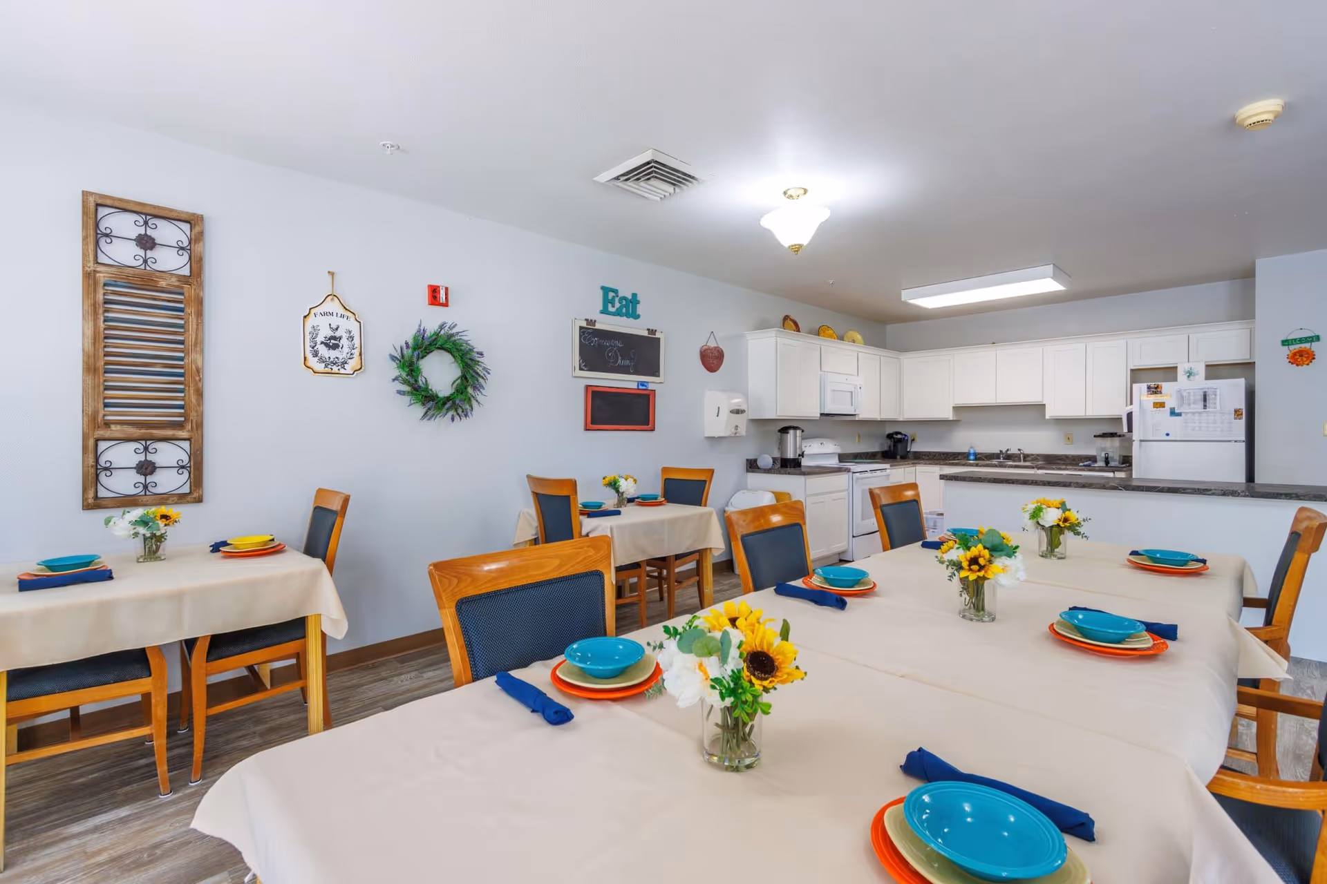 A dining area with several tables covered in beige tablecloths, each set with colorful plates, bowls, and blue napkins. Small flower arrangements with sunflowers and white flowers are placed on the tables. The room has wooden chairs with blue cushions and a kitchen area with white cabinets, a refrigerator, stove, and microwave in the background. Wall decorations include a wreath, a sign that says 'Eat', and other rustic decor.