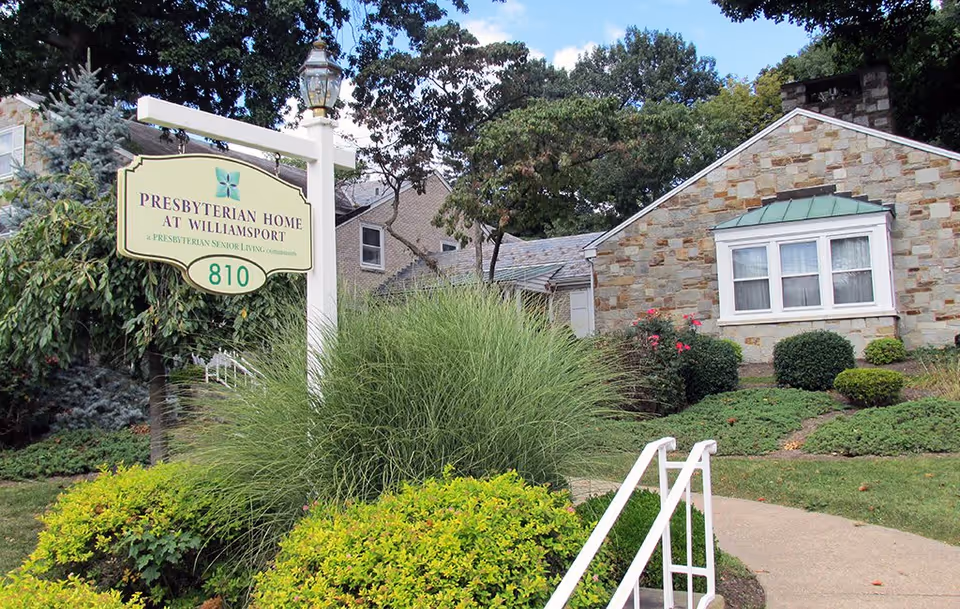 Exterior view of Presbyterian Home at Williamsport showing a stone building with white-framed windows, surrounded by green bushes and trees. A white post sign with the facility name and address number 810 is prominently displayed near the sidewalk with a white handrail leading up to the entrance.