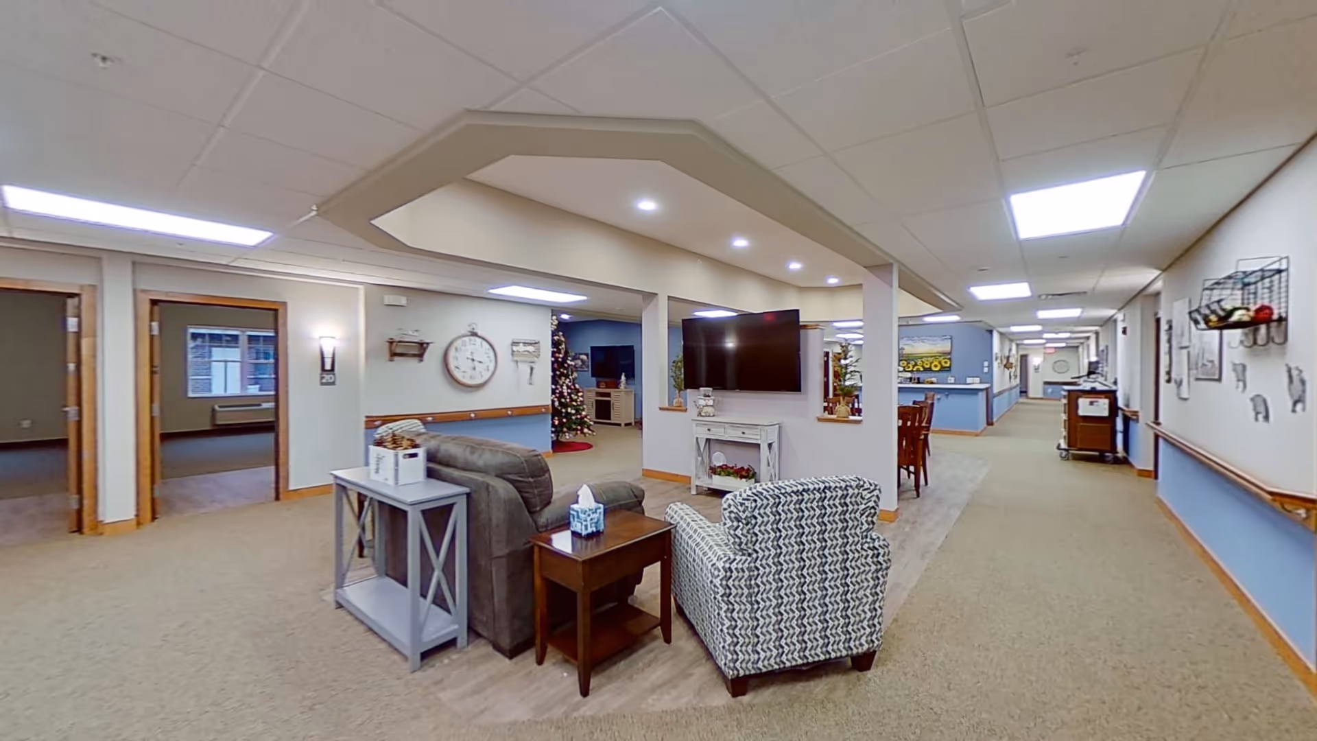 Interior view of a senior living facility common area with a seating arrangement including a patterned armchair and a gray sofa facing a wall-mounted TV. The space is well-lit with ceiling lights and decorated with a large wall clock, a Christmas tree, and various wall art. There is a long hallway extending to the right with handrails and additional seating and decor visible.