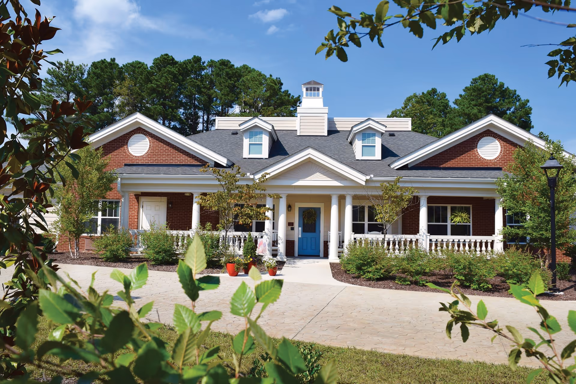 Front exterior view of a single-story brick building with white columns, a blue front door, and a porch surrounded by greenery and trees under a clear blue sky.