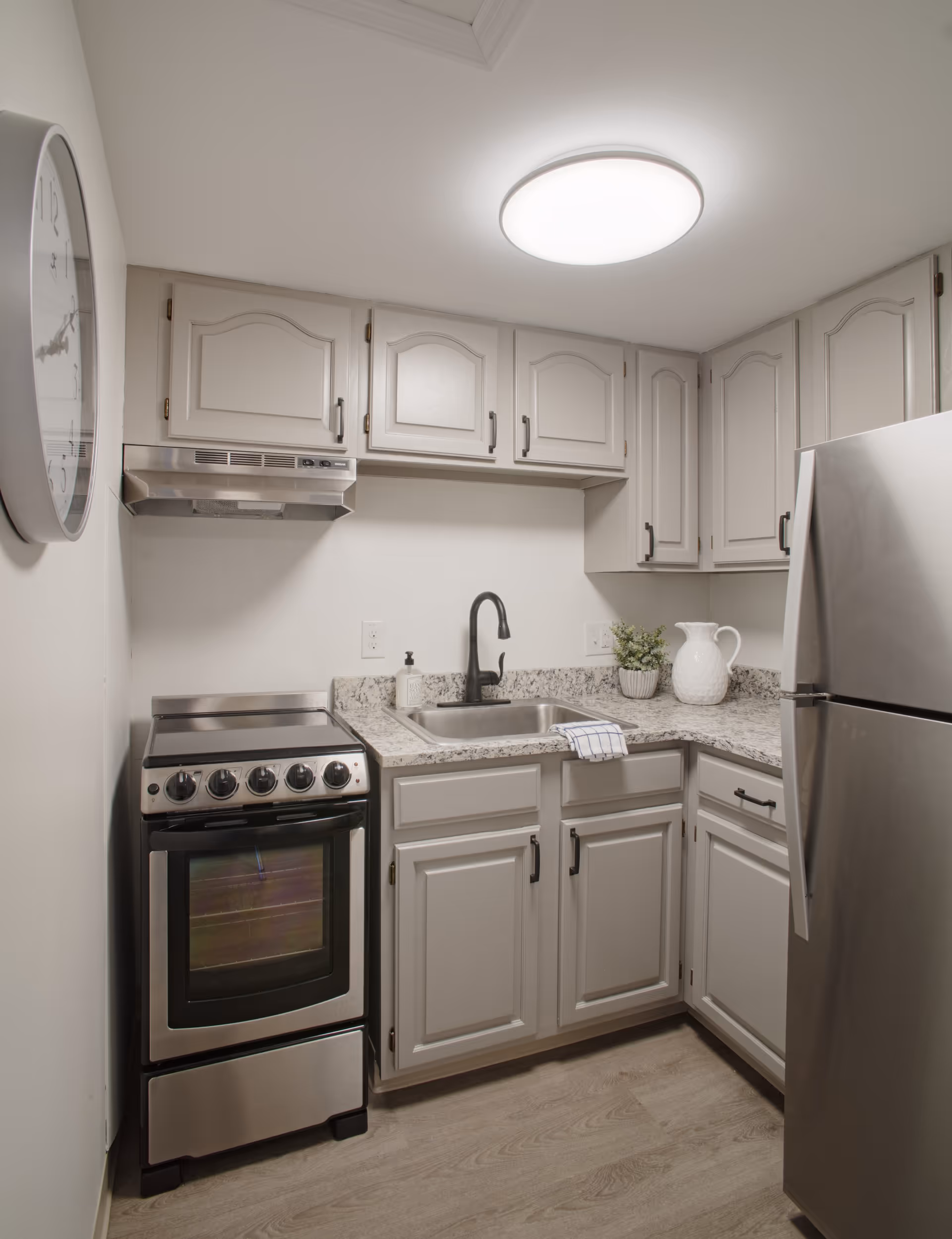A compact kitchen featuring light gray cabinets with black handles, a stainless steel stove and refrigerator, a granite countertop with a built-in sink and black faucet. A white pitcher and a small potted plant are on the counter, and a round wall clock is visible on the left wall.