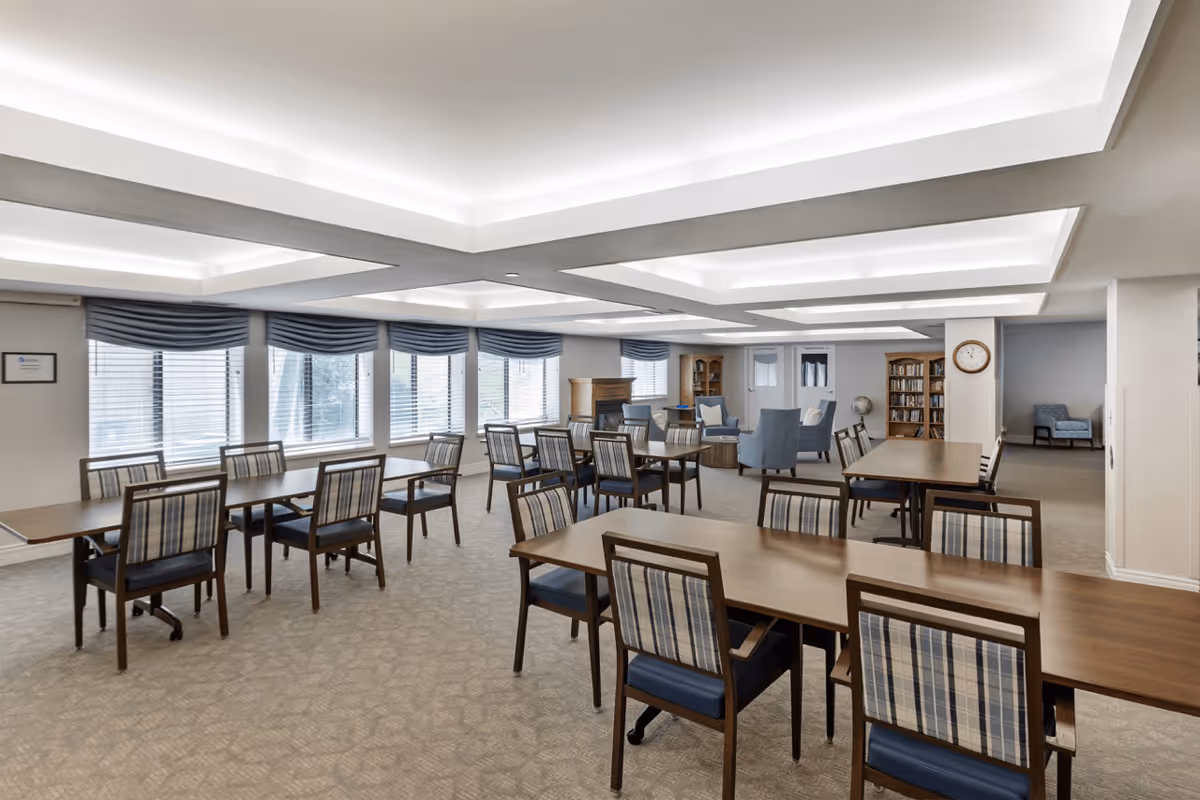A spacious senior living common area with multiple wooden tables and chairs arranged neatly. The room features large windows with blue valances, a carpeted floor, and a ceiling with recessed lighting. In the background, there are comfortable armchairs, a fireplace, bookshelves, and a wall clock.