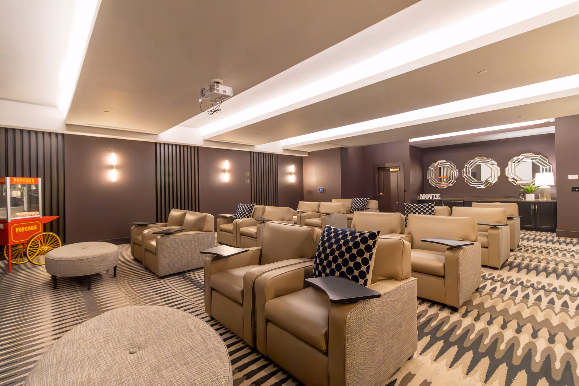 A cozy screening room with rows of leather recliners, a ceiling-mounted projector, patterned carpet, and a popcorn cart.