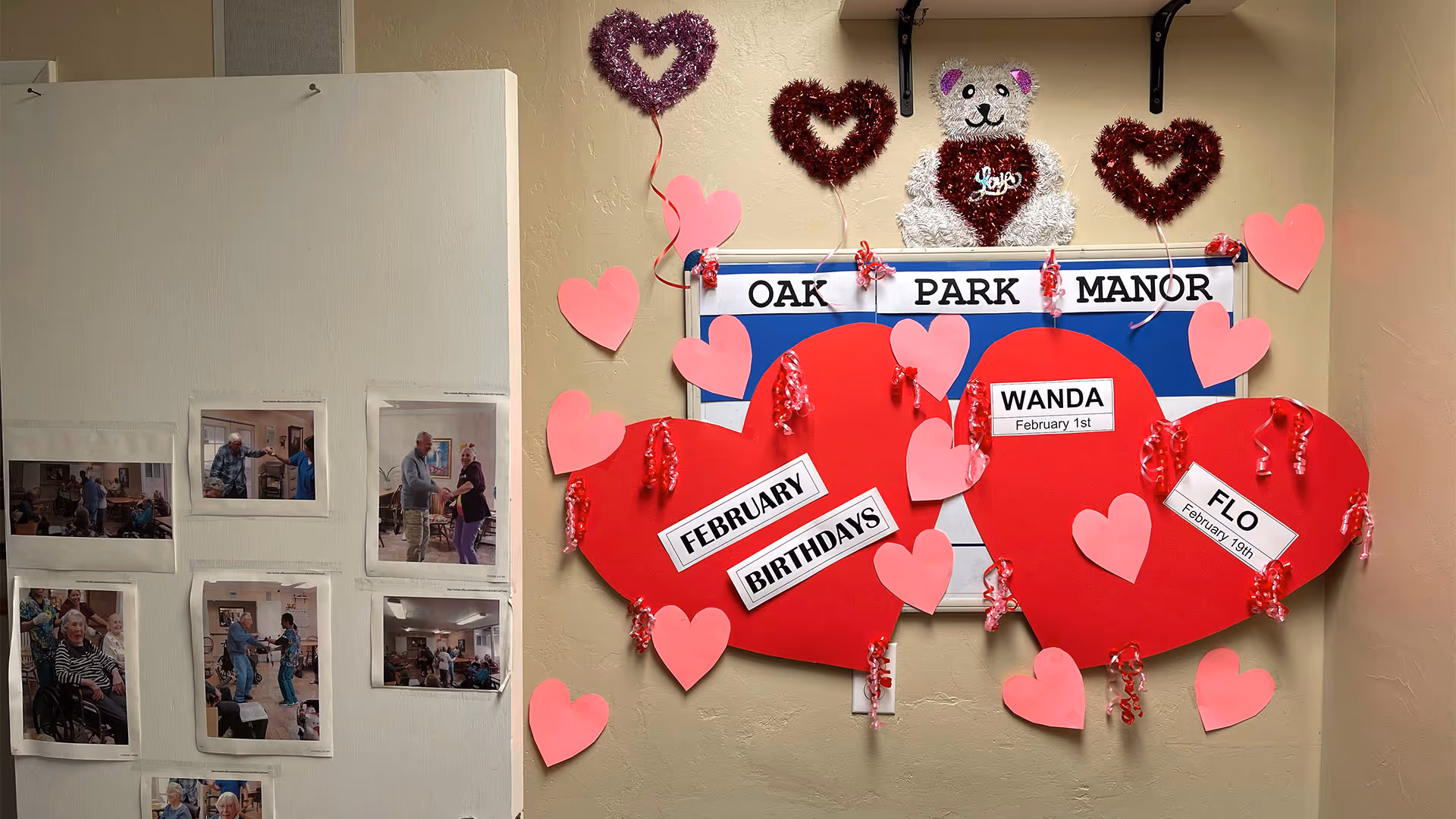 A bulletin board decorated with Valentine-themed decorations including red and pink paper hearts, a teddy bear made of tinsel, and a sign that reads 'OAK PARK MANOR'. Two large red hearts display February birthdays for Wanda on February 1st and Flo on February 19th. To the left, there are several photos of elderly residents engaging in activities.