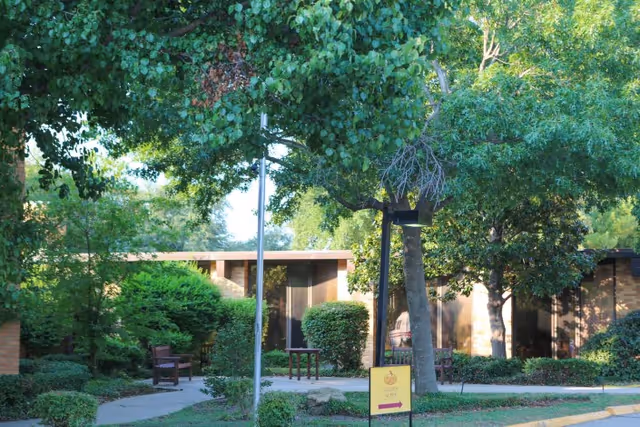 Outdoor view of a senior living facility with a pathway surrounded by green trees and bushes. There are benches along the path and a yellow sign with an arrow pointing to the right. The building is partially visible in the background with large windows.