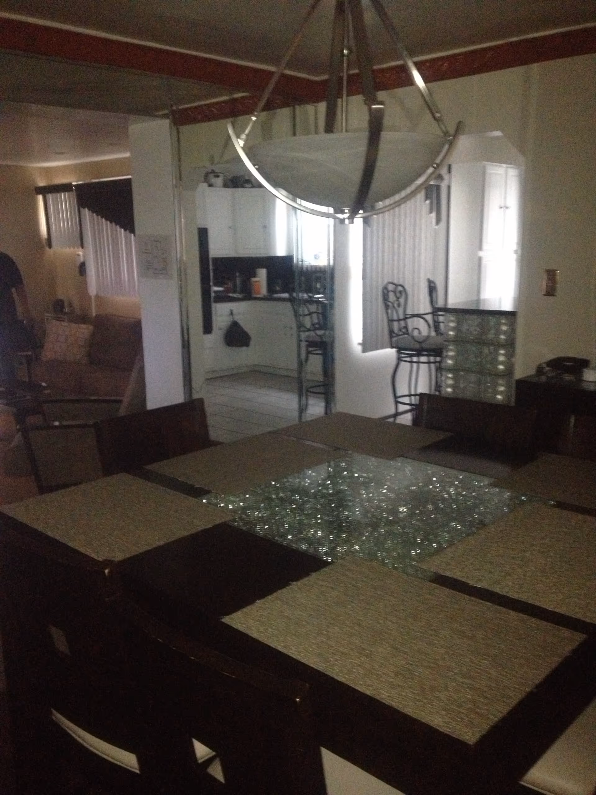 Dining room table with placemats and a chandelier in an open interior showing an adjacent kitchen and living area.