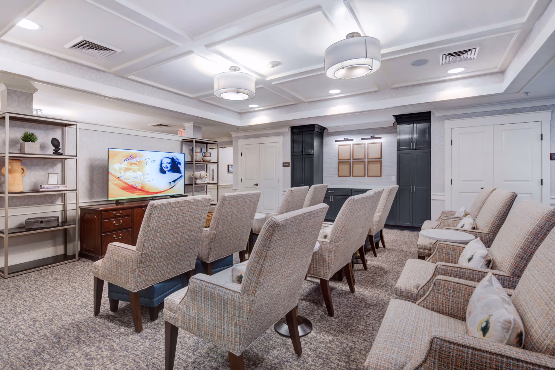 A well-lit communal media room with rows of upholstered chairs facing a flatscreen TV, shelving, and built-in cabinets.