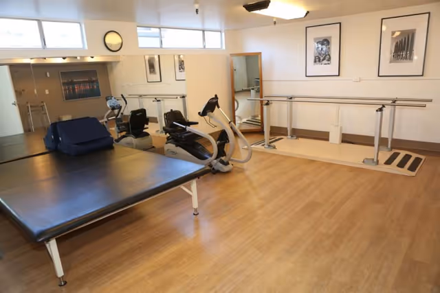 A therapy or rehabilitation room with a large padded table, exercise equipment including a recumbent bike, parallel bars for walking support, mirrors on the walls, framed pictures, and a clock above the windows.