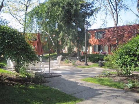 Outdoor courtyard area with paved walkways, green bushes, trees, and patio chairs. A brick building with windows is visible in the background under a clear blue sky.