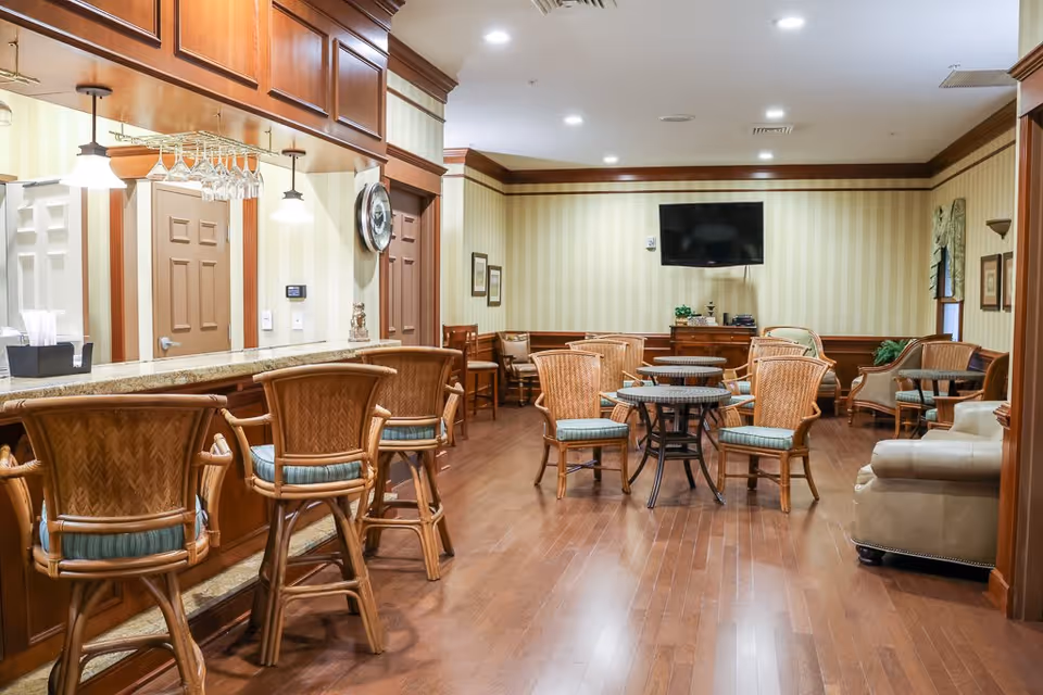 A cozy senior living common area with wooden flooring, wicker chairs with cushions around small round tables, a bar counter with high chairs, and a wall-mounted flat screen TV. The room has warm lighting, wood paneling, and striped wallpaper.