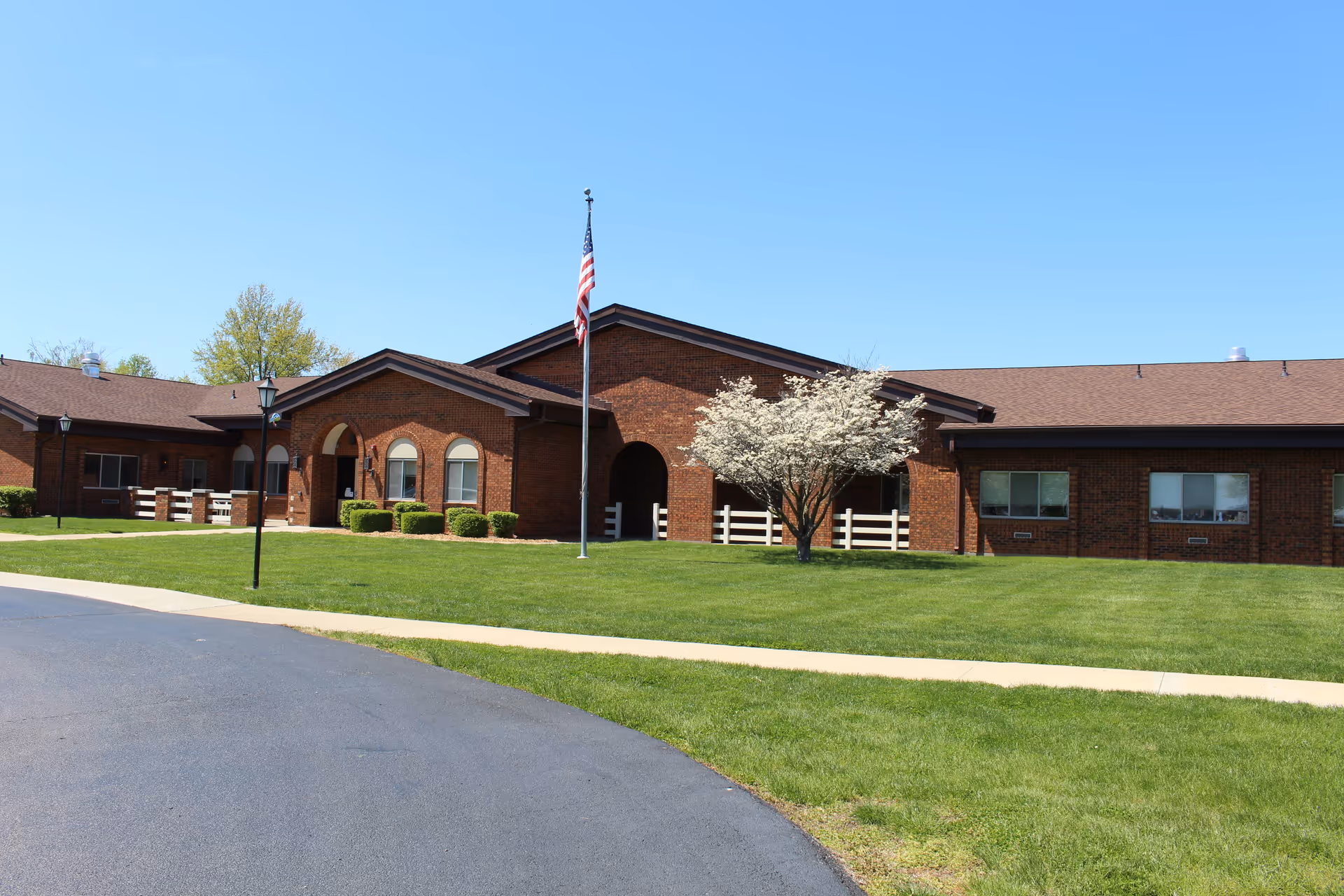 Exterior view of a single-story brick building with a brown roof, an American flag on a flagpole, a blooming tree, and well-maintained green lawn under a clear blue sky.