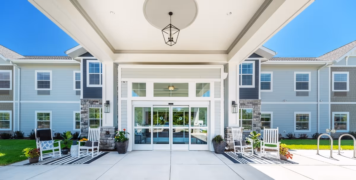 Entrance of a senior living facility with a covered porch area featuring white rocking chairs on striped rugs, potted plants, and automatic sliding glass doors leading inside. The building exterior is light-colored with multiple windows and stone accents under a clear blue sky.