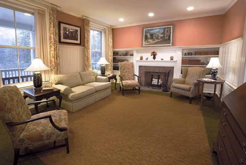 A cozy living room with a beige sofa, three upholstered armchairs, two side tables with lamps, a fireplace with a painting above it, and windows with floral curtains letting in natural light.